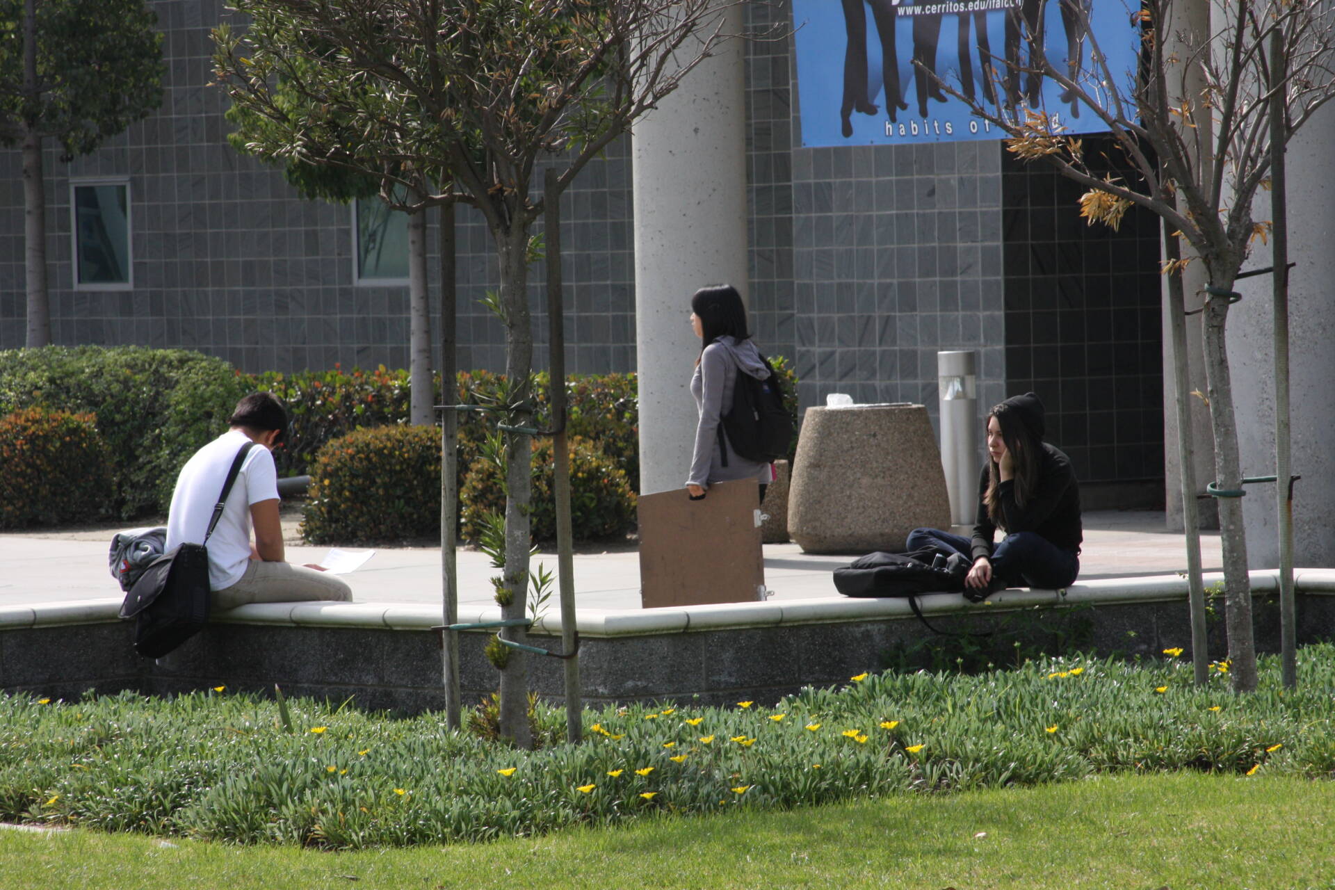 Two Cerritos College students, a young man and a young woman, sit on a low stone wall outside a gray classroom building. A third woman is passing by them on the sidewalk, carrying an artist's portfolio.